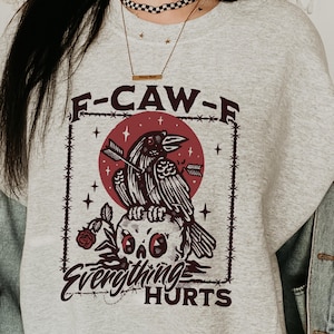 May include: A light gray sweatshirt with a graphic design featuring a crow perched on a skull, pierced by an arrow, with the text "F-CAW-F" and "Everything Hurts". The design is in shades of red and black.