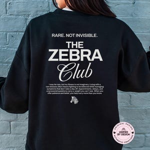 May include: Black sweatshirt with white text that reads "RARE. NOT INVISIBLE. THE ZEBRA Club." Additional text below describes living with a rare disease. A small zebra graphic is also present. The sweatshirt is a crew neck.