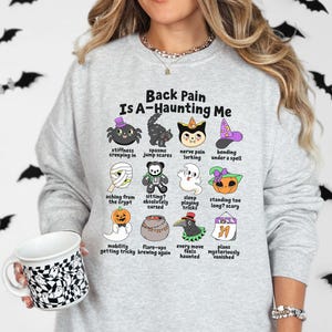 May include: A light gray crewneck sweatshirt featuring a graphic with the text Back Pain Is A-Haunting Me and twelve cartoon illustrations of Halloween characters with labels describing physical ailments, held with a black and white ghost mug.