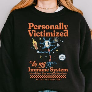 May include: Black sweatshirt featuring the text "Personally Victimized by my Immune System" in orange and white. The design includes a cartoon neuron with a flame and lightning bolts. The words "Multiple Sclerosis Club" are also present.
