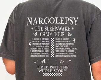 Może przedstawiać: Ciemnoszary t-shirt z krótkim rękawem z białym napisem na plecach o treści NARCOLEPSY THE SLEEP-WAKE CHAOS TOUR, wraz z listą objawów, ikonami motyli, błyskawicami oraz frazą TIRED ISN'T THE WHOLE STORY.
