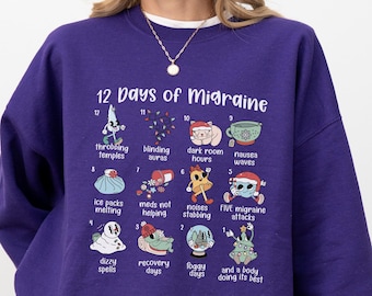 May include: A deep purple long-sleeve sweatshirt featuring a graphic titled 12 Days of Migraine. The design includes twelve numbered illustrations with text labels describing various symptoms and experiences related to migraine pain and recovery.