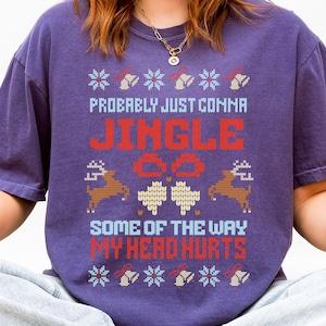 Puede incluir: Una camiseta morada con el texto "Probably Just Comma Jingle All The Way My Head Hurts" en rojo y azul. La camiseta tiene un diseño festivo con renos y patrones de copos de nieve, ideal para la temporada navideña.