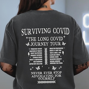 May include: A dark gray t-shirt with white text that reads "Surviving COVID + The Long COVID + Journey Tour" and lists symptoms of long COVID. The text also says "Never ever stop advocating for yourself".