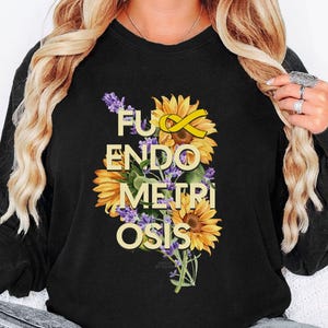 Peut inclure: Sweat-shirt noir avec un motif floral composé de tournesols, de lavande et d'un ruban jaune. Le texte "F*ck Endometriosis" est en or. Le motif est centré sur le devant du haut à manches longues.