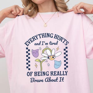 Könnte beinhalten: Hellrosa T-Shirt mit dem Text "EVERYTHING HURTS and I'm tired OF BEING REALLY Brave About It". Das Design zeigt eine Gänseblümchen mit Gesicht, Blitzen und Cartoon-Figuren.