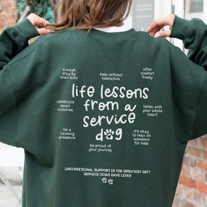 Może przedstawiać: Zielony sweter z białym tekstem, który brzmi "Life lessons from a service dog" z odciskami łap i innymi inspirującymi frazami.
