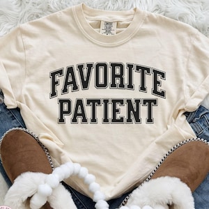 Puede incluir: Una camiseta de manga corta color crema con el texto negro FAVORITE PATIENT impreso en una fuente universitaria arqueada y llamativa en el pecho. La camiseta se coloca sobre unos vaqueros azules y zapatillas de ante marrón con borde de piel sintética.