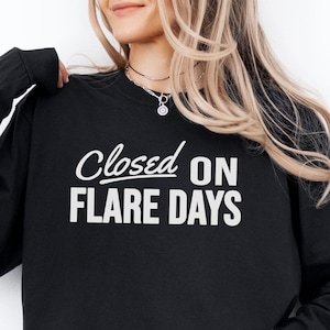 Puede incluir: Una sudadera negra de manga larga con el texto blanco "Closed ON FLARE DAYS" impreso en el pecho. La prenda tiene cuello redondo y mangas largas con puños acanalados. Se observa un pequeño logotipo circular con el texto "UNIFIED BY DESIGN".