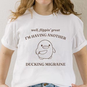 May include: White t-shirt with brown text that reads "well, flippin' great I'M HAVING ANOTHER DUCKING MIGRAINE". A cartoon penguin with a knife is pictured below the text.