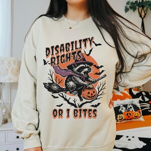 Puede incluir: Sudadera crema con un gráfico de Halloween. El diseño presenta un mapache disfrazado de bruja montado en una escoba, sosteniendo una calabaza y el texto "DISABILITY RIGHTS OR I BITES" en naranja y negro.