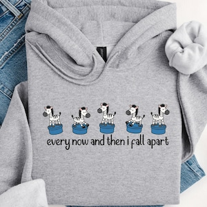 Puede incluir: Una sudadera con capucha gris jaspeado con un gráfico de cinco cebras sobre plataformas azules. Las cebras tienen rayas blancas y negras y llevan sombreros. El texto "every now and then I fall apart" está debajo.