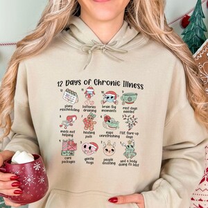 May include: A beige hooded sweatshirt featuring a graphic titled 12 Days of Chronic Illness. The design includes twelve numbered illustrations with text labels such as plans rescheduling, batteries draining, brain fog, and heating pads.