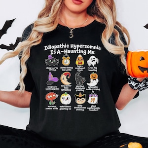 May include: A black t-shirt featuring the text Idiopathic Hypersomnia Is A-Haunting Me above a grid of twelve Halloween-themed illustrations with captions including a spider, alarm clock, candy corn, ghost, witch hat, moon, cat, pumpkin, brain, and calendar.