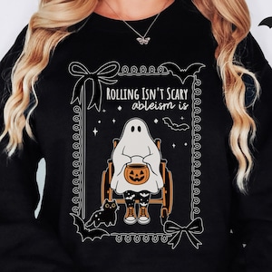 May include: Black sweatshirt with a Halloween-themed graphic. The design features a ghost in a wheelchair holding a jack-o'-lantern, surrounded by bats, stars, and the text "Rolling isn't scary, ableism is."