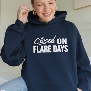Puede incluir: Una sudadera con capucha de color azul marino que presenta el texto blanco "Closed ON FLARE DAYS" impreso en el pecho. La prenda incluye una capucha con cordones, mangas largas con puños acanalados y un bolsillo canguro frontal.