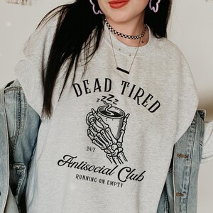 Puede incluir: Sudadera gris con un gráfico negro de una mano esquelética sosteniendo una taza de café. El texto "Dead Tired" está por encima del gráfico y "Antisocial Club" está por debajo del gráfico. El texto "24/7 Running on Empty" está debajo del nombre del club.