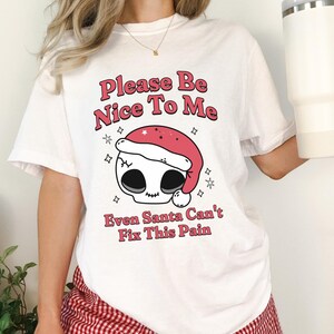 Puede incluir: Camiseta blanca con el texto "Please Be Nice To Me" sobre una calavera con un gorro de Papá Noel. La camiseta también tiene el texto "Even Santa Can't Fix This Pain". La modelo lleva un pantalón corto a cuadros rojos y blancos.