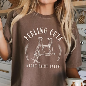 May include: A brown t-shirt with the text "FEELING CUTE MIGHT FAINT LATER" in a white, circular design. The design includes a goat illustration. The person is holding a mug with bear illustrations.