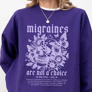 Könnte beinhalten: Ein lila Sweatshirt mit dem weißen Schriftzug "migraines are not a choice" in gotischer Schrift. Eine Totenkopf-Grafik mit floralen und Schmetterlingsakzenten ist unter dem Text zentriert. Der Satz "AND STILL I RISE" ist ebenfalls aufgedruckt.