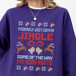 Puede incluir: Una sudadera morada con un diseño festivo. El texto dice "Probably Just Comma Jingle" en rojo y blanco, con gráficos de renos y copos de nieve. También se muestra la frase "Some of the way my head hurts".