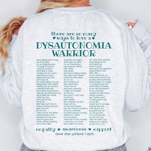 Puede incluir: Sudadera gris claro con el texto "there are so many ways to love a Dysautonomia Warrior" en verde azulado. La parte posterior de la camisa enumera frases de apoyo. La parte inferior de la camisa tiene las palabras "empathy, awareness, support" y "love me where I am".