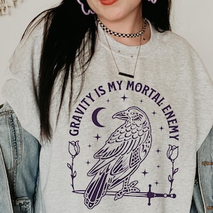 Puede incluir: Sudadera gris claro con un diseño morado de un cuervo, luna creciente, estrellas y el texto "GRAVITY IS MY MORTAL ENEMY". El cuervo está posado sobre una espada, enmarcado por elementos florales.