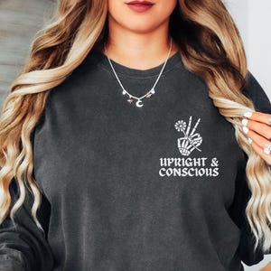May include: A dark grey sweatshirt with a white graphic of a skeleton hand making a peace sign with a flower. The text "Upright & Conscious" is below the graphic.