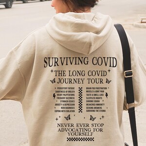 Puede incluir: Sudadera beige con un estampado gráfico negro que dice "Surviving COVID + The Long COVID + Journey Tour" y enumera los síntomas del COVID largo. El gráfico también incluye el texto "Never Ever Stop Advocating For Yourself".