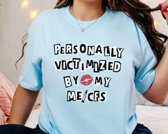 May include: Light blue t-shirt with the text "PERSONALLY VICTIMIZED BY MY ME/CFS" in a black, blocky font. A red lipstick kiss graphic is between "BY" and "MY". The shirt is a crew neck and short-sleeved.