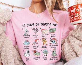 Puede incluir: Una camiseta rosa claro con un gráfico titulado 12 Days of Migraine que muestra doce ilustraciones numeradas de varios síntomas y remedios, incluyendo sienes palpitantes, auras cegadoras, horas en cuartos oscuros, náuseas y un árbol de Navidad.