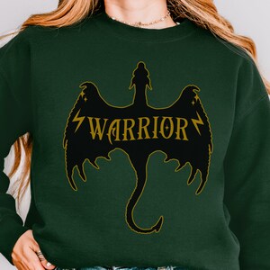 May include: Dark green sweatshirt with a black dragon design and gold lettering that reads "WARRIOR". The dragon has outstretched wings and a curved tail. The sweatshirt is a plain color.