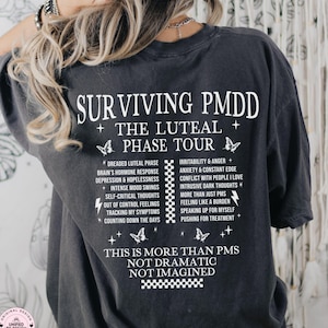 Könnte beinhalten: Dunkelgraues T-Shirt mit weißem Aufdruck "SURVIVING PMDD" und "THE LUTEAL PHASE TOUR". Das Shirt listet verschiedene Symptome und Phrasen im Zusammenhang mit PMDD auf. Das Wort "PEPPER" ist unten aufgedruckt.