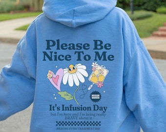 May include: Light blue hoodie with a graphic of a daisy, a heart, and a medical bag. The text on the hoodie reads "Please Be Nice To Me", "It's Infusion Day", "but I'm here and I'm being really BRAVE about it", and "BRAVING EVERY TREATMENT DAY".