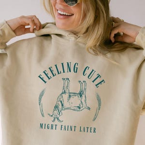 May include: Beige hoodie with a hood and a kangaroo pocket. The front features a teal graphic of a goat and the text "FEELING CUTE MIGHT FAINT LATER". The hoodie is worn by a person wearing a beige cap and sunglasses.