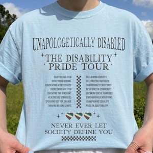 May include: Light blue t-shirt with the text "UNAPOLOGETICALLY DISABLED THE DISABILITY PRIDE TOUR". The shirt features a list of phrases and a graphic of three hearts. The text "NEVER EVER LET SOCIETY DEFINE YOU" is also printed on the shirt.