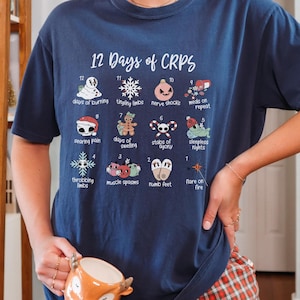 Op de afbeelding: Een marineblauw T-shirt met een witte afbeelding van de "12 dagen van CRPS" met een kerstthema. De afbeelding toont 12 verschillende symptomen van CRPS, elk met een bijbehorend nummer en een feestelijk pictogram.