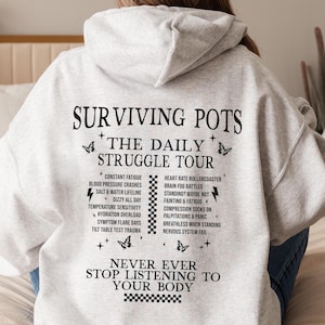 Puede incluir: Sudadera gris con capucha con texto negro que dice "Surviving Pots + The Daily Struggle Tour" y enumera los síntomas del POTS, una condición que afecta el sistema nervioso autónomo. El texto también dice "Never Ever Stop Listening To Your Body".