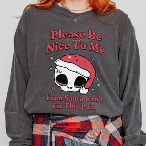Puede incluir: Camiseta de manga larga gris oscuro con el texto "Please Be Nice To Me" y "Even Santa Can't Fix This Pain". La camiseta presenta una calavera con un gorro de Papá Noel. La camiseta está hecha de un material suave y cómodo.