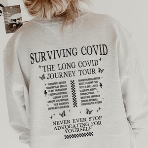 Puede incluir: Sudadera blanca con texto negro que dice "Surviving COVID + The Long COVID + Journey Tour" y enumera los síntomas del COVID largo. El texto también dice "Never Ever Stop Advocating For Yourself".