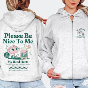 Puede incluir: Sudadera gris con cremallera y capucha, con un gráfico verde y blanco en la parte posterior que dice "Please Be Nice To Me" y un cerebro de dibujos animados con un frasco de píldoras. La parte delantera de la sudadera tiene un pequeño gráfico verde y blanco que dice "Headache Survivor".