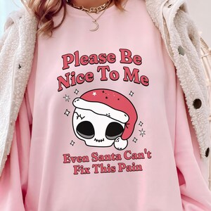 Puede incluir: Sudadera rosa claro con el texto "Please Be Nice To Me" y "Even Santa Can't Fix This Pain". El diseño presenta una calavera con un gorro de Papá Noel, rodeada de estrellas. Un diseño festivo y cómodo.