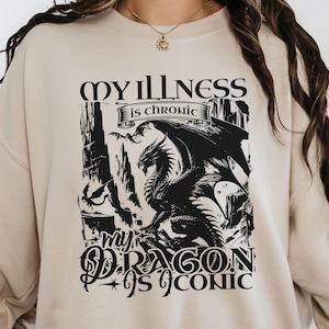May include: Beige sweatshirt featuring a black dragon graphic and the text "MY ILLNESS is chronic, my DRAGON is iconic". The design includes a detailed dragon illustration and gothic lettering.