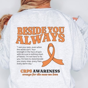 May include: Pale grey long-sleeved sweatshirt featuring the orange text "BESIDE YOU ALWAYS". An orange ribbon graphic and a quote are below the text. The words "CRPS AWARENESS" and "orange for the ones we love" are also printed on the sweatshirt.