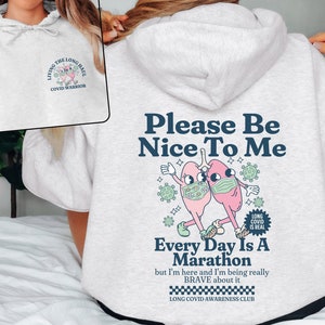 Puede incluir: Sudadera gris con un gráfico de dos pulmones rosas con mascarillas y el texto "Please Be Nice To Me" y "Every Day Is A Marathon". El gráfico también incluye el texto "Long Covid Is Real" y "Long Covid Awareness Club".