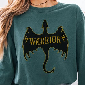 May include: Teal sweatshirt with a black dragon design and gold "WARRIOR" text. The dragon has lightning bolt details. The long-sleeved top has a crew neck. The image shows a close-up of the upper body.