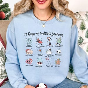 Op de afbeelding: Lichtblauw sweatshirt met de tekst "12 Days of Multiple Sclerosis" en illustraties van verschillende symptomen. Het sweatshirt is lichtblauw en heeft een ronde hals.