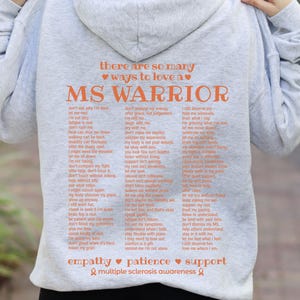 May include: A light gray hoodie with the words "MS WARRIOR" in orange, along with a list of supportive phrases. The hoodie also features the words "empathy, patience, support" and the multiple sclerosis awareness ribbon.