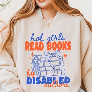 Op de afbeelding: Een beige sweater met ronde hals en een grafische print met de tekst "hot girls READ BOOKS by DISABLED authors". Het ontwerp bevat een stapel van vier boeken die zijn samengebonden met een lint in blauw en oranje, met kleine sterren.