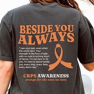 Op de afbeelding: Donkergrijs T-shirt met de oranje tekst "BESIDE YOU ALWAYS". Daaronder een citaat en een oranje lintafbeelding. De tekst "CRPS AWARENESS orange for the ones we love" staat ook op het shirt.